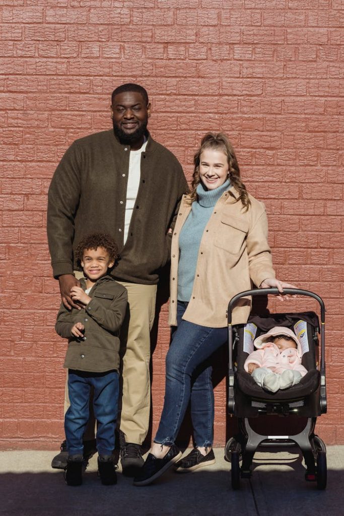 A joyful family portrait with parents, child, and baby stroller in sunlight.
