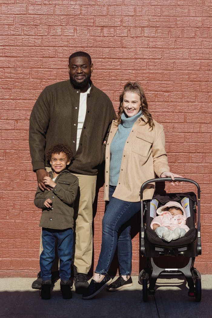 A joyful family portrait with parents, child, and baby stroller in sunlight.